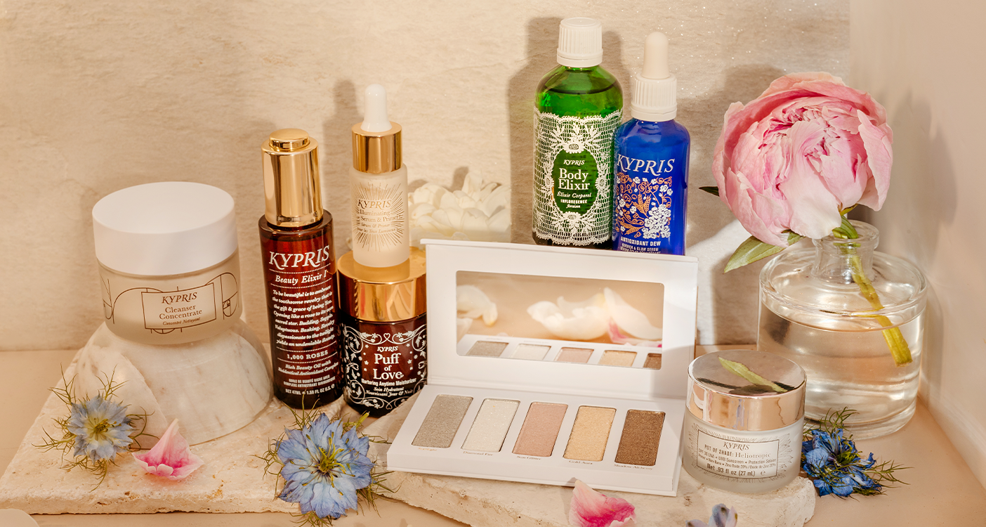 Product shot of Kypris Ad Astra, Cleanser Concentrate, Antioxidant Dew, Body Elixir, Puff of Love, Illuminating Eye Shadow & Shimmer Palette against a stone and floral background.