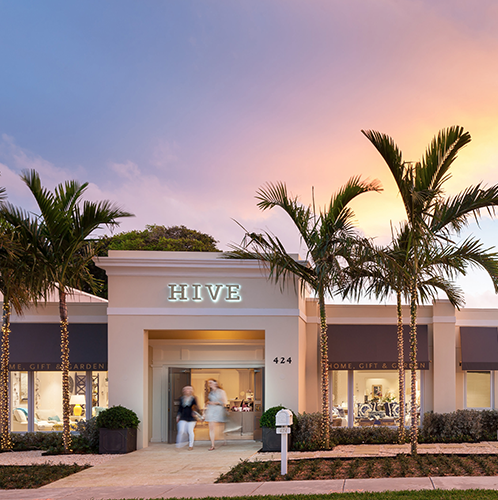 Hive Palm Beach store front photo in West Palm Beach, Florida. A cream building with a neon sign of their logo, with palm tree landscaping and a beautiful sunset sky.