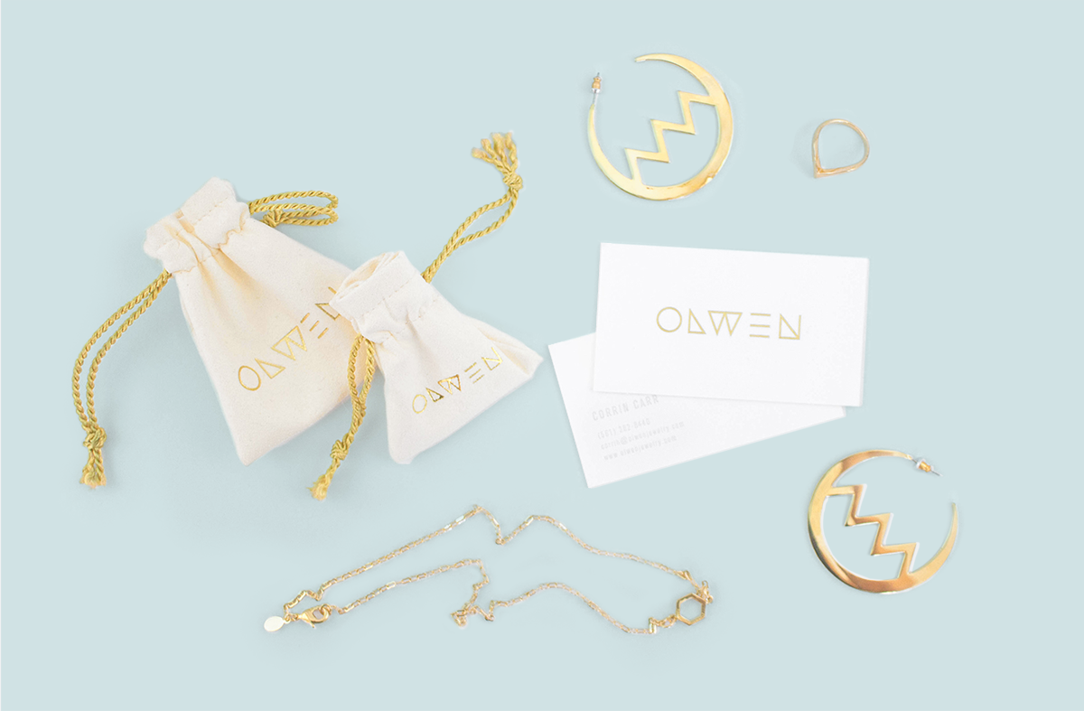 Cream colored cloth jewelry bags with golden drawstrings and OLWEN logo, next to white business cards with gold foiled logo, zigzag hoop earrings, a ring and necklace against a baby blue background.