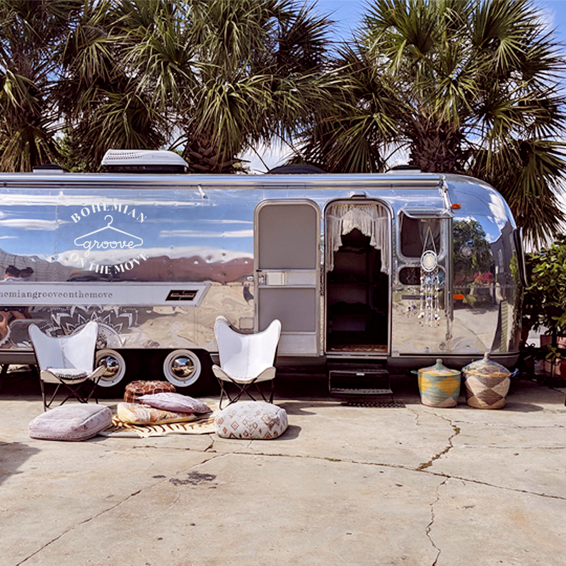 Two white chairs sit in front of an iconic "silver bullet" airstream, featuring a logo reading "Bohemian Groove on the Move" with palm trees in the background