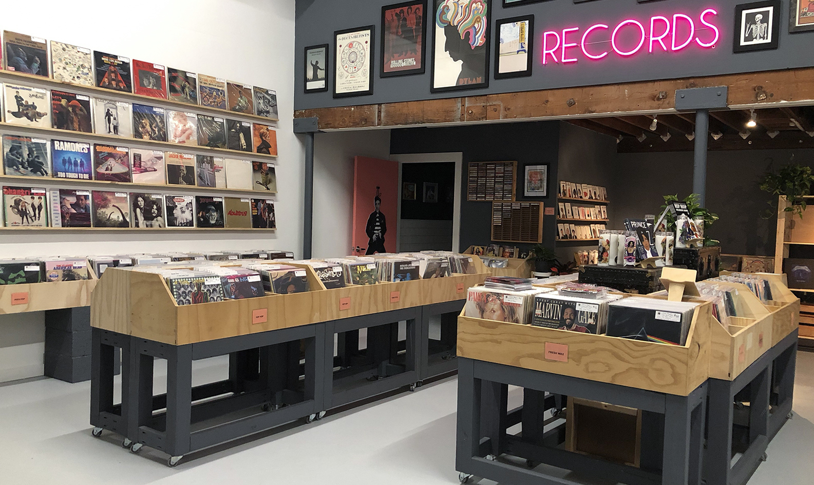West Palm Beach's local record shop Rust & Wax interior with aisles of record bins. The wall above features a collection of vintage music posters and a bright pink neon sign of the word "records"