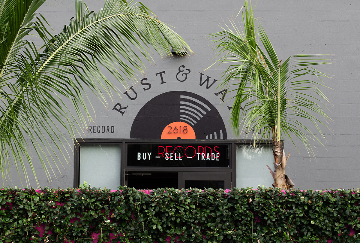 Hand painted mural signage for Rust & Wax record shop in West Palm Beach, featuring a large illustrated record with an orange label. The logo is on a curve above, with the type Record Store flanking the sides of the record, & Buy Sell Trade is below.