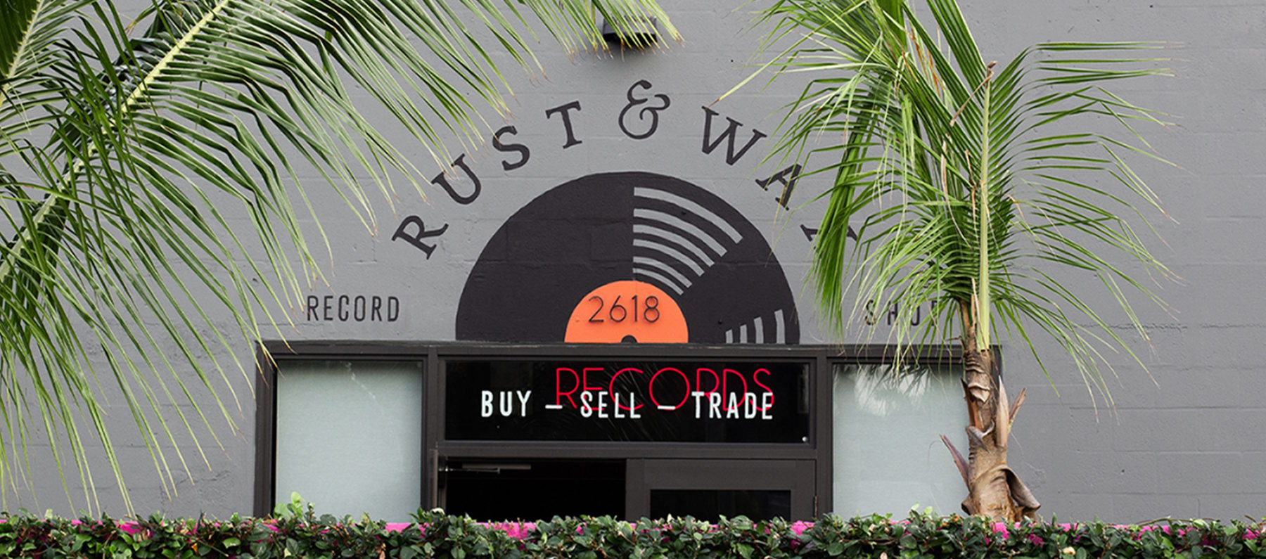 Hand painted mural signage for Rust & Wax record shop in West Palm Beach, featuring a large illustrated record with an orange label. The logo is on a curve above, with the type Record Store flanking the sides of the record, & Buy Sell Trade is below.