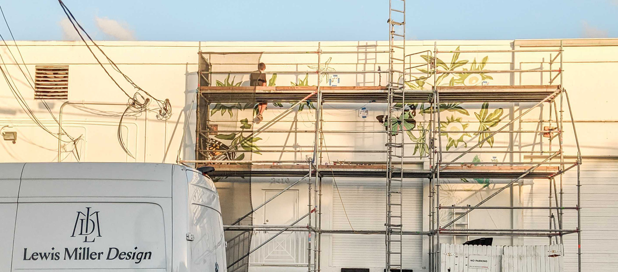 Process shot of painters from Gather & Seek on scaffolding, in front of the Lewis Miller Design building, painting a mural of passion flowers, leaves and vines, and butterflies.