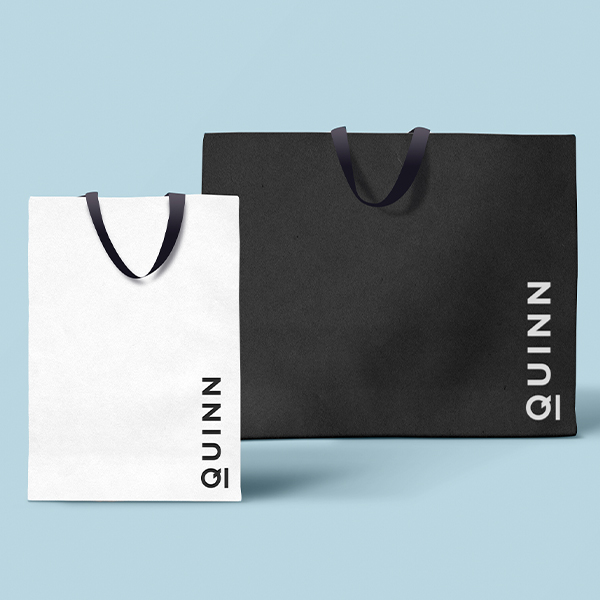 Black and white shopping bag mockups with a vertical logo in the lower right hand corner of the bags in the inverted color, on a light blue background.