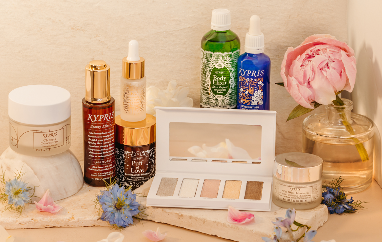 KYPRIS Cleanser Concentrate, Beauty Elixir I, Puff of Love, Illuminating Eye Serum & Primer, Shimmer Palette, Boxy Elixir, Antioxidant Dew, and Pot of Shade: Heliotropic placed on a creamy travertine stone tiles with flowers scattered around.
