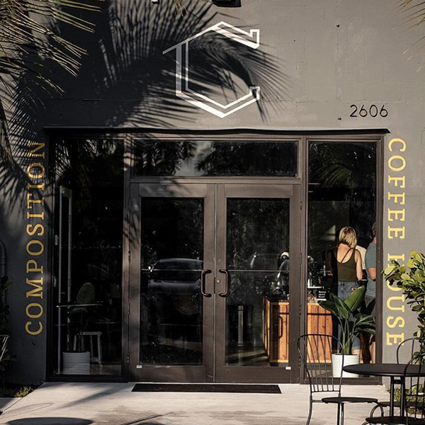 Composition Coffee storefront location in Industry Alley of West Palm Beach. A gray building with black aluminum and glass doors, a large C Icon appears above the door, with the words Composition and Coffee House vertically along the windows.